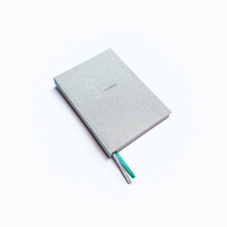 Jeem Journal standing upright - a sleek, silver fabric cover and two bookmark ribbons (one green and one silver)